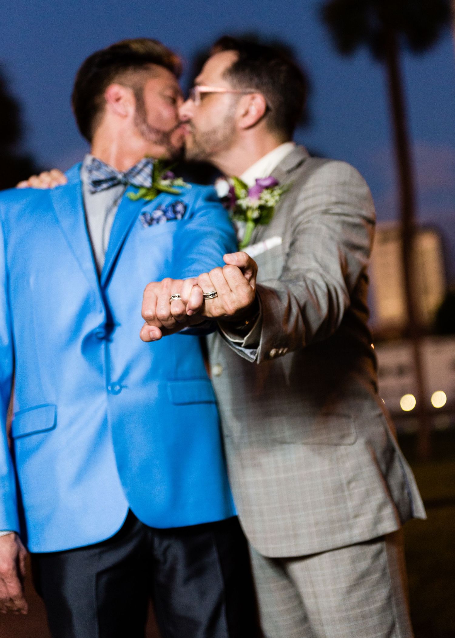 Two grooms kissing and showing wedding rings at night, one in blue suit, one in gray suit.