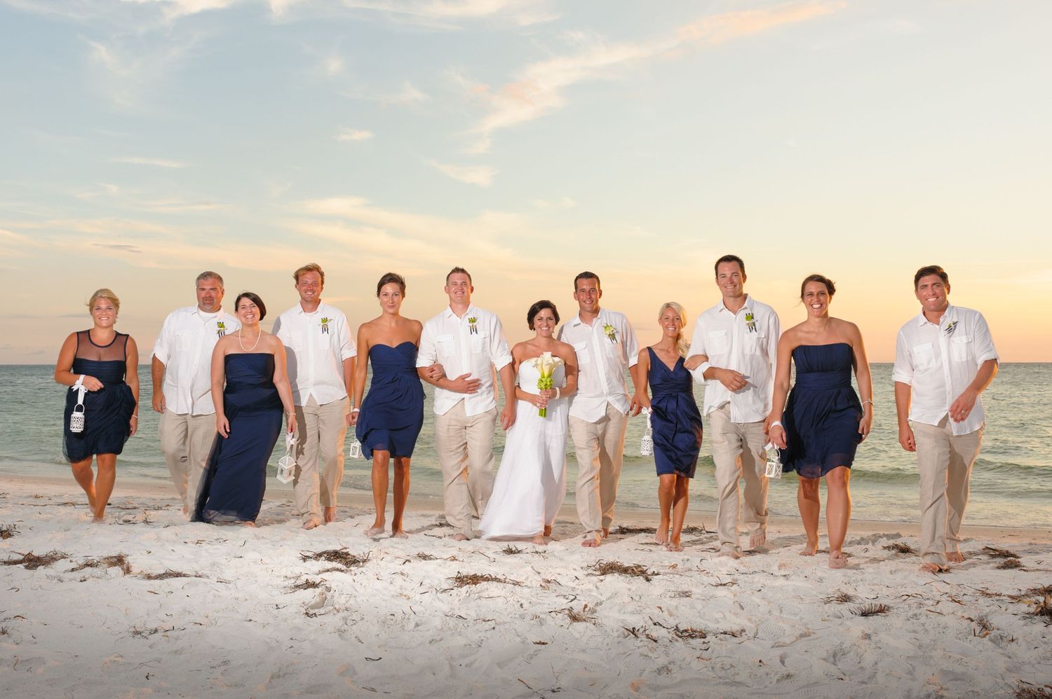 Wedding party posing on a sandy beach at sunset, bridesmaids in navy dresses and groomsmen in tan suits.