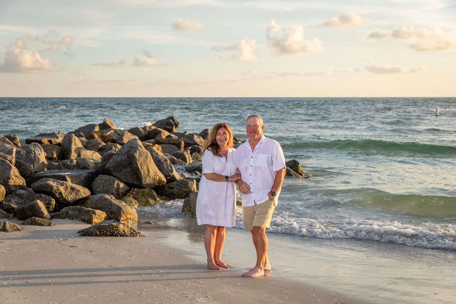 Couple smiling together on a sandy beach near rocky jetty with ocean waves and golden sunset sky.