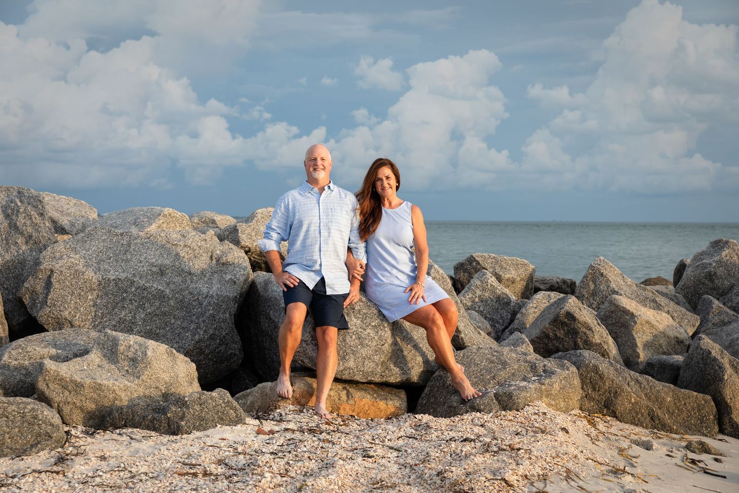 Couple smiling on rocky beach with ocean and cloudy sky backdrop on a sunny day.