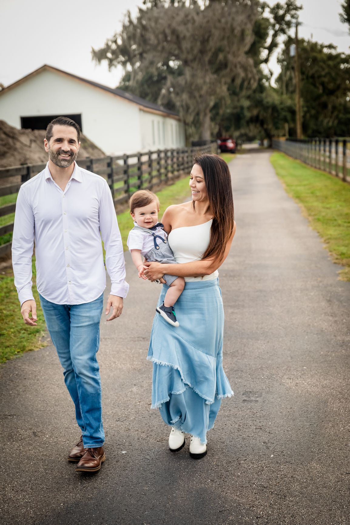 Happy young family with baby boy walking along a scenic farm pathway surrounded by wooden fences.