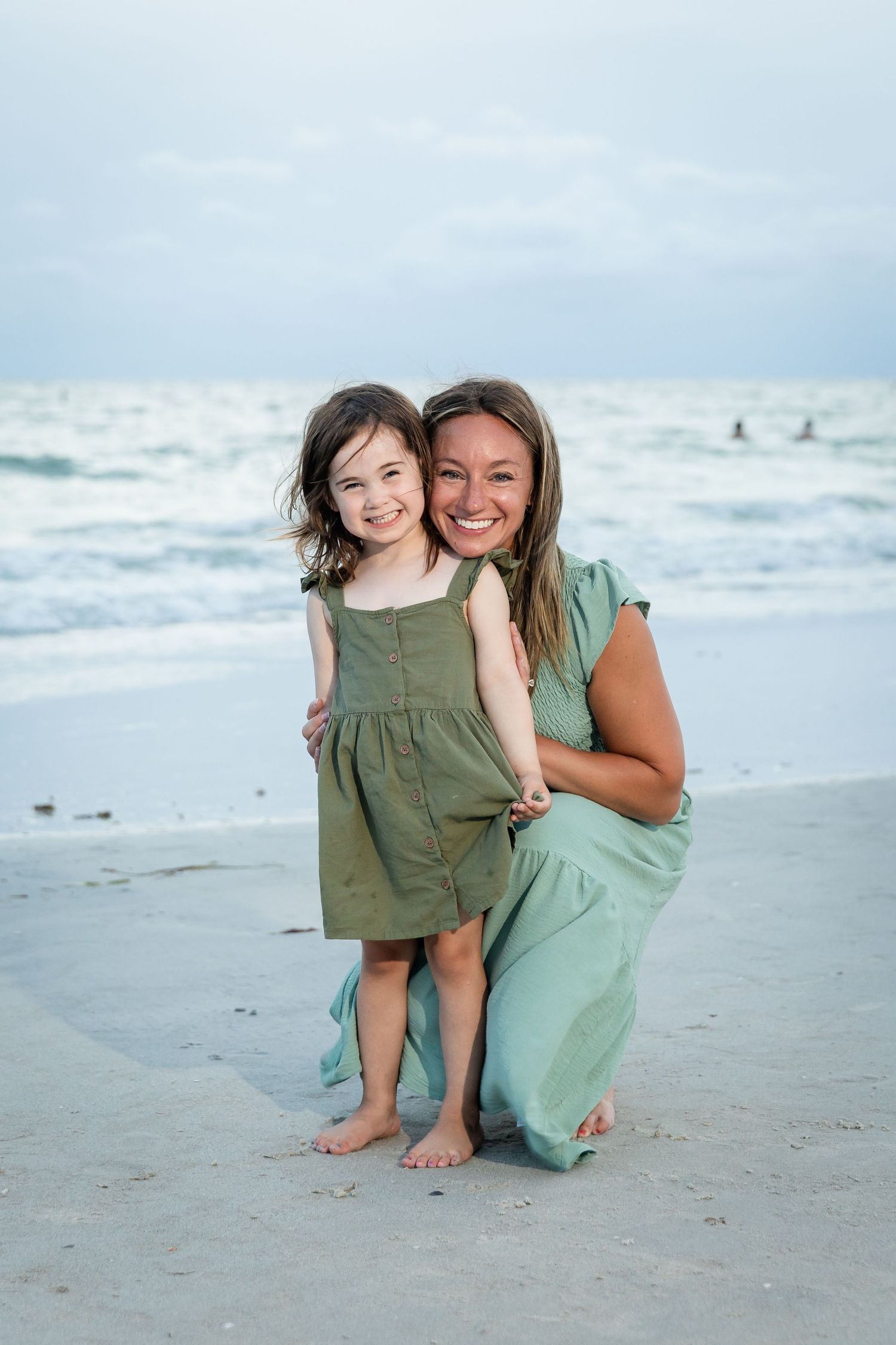 Smiling mother in mint dress crouching beside young daughter in green dress on beach.