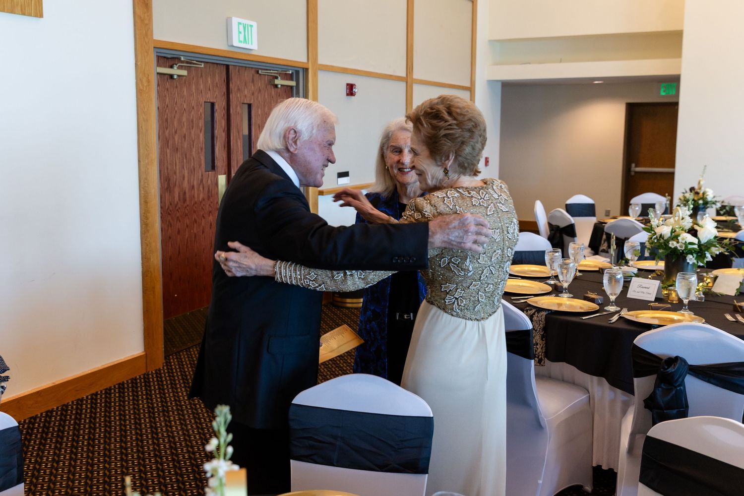 Elderly couple sharing a warm embrace at an elegant wedding reception with black and white table settings.
