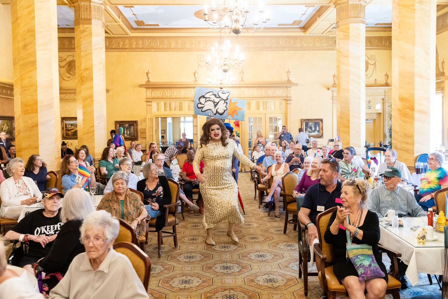 A drag performer walks the runway in a golden gown at a packed elegant ballroom event with diverse seated audience.