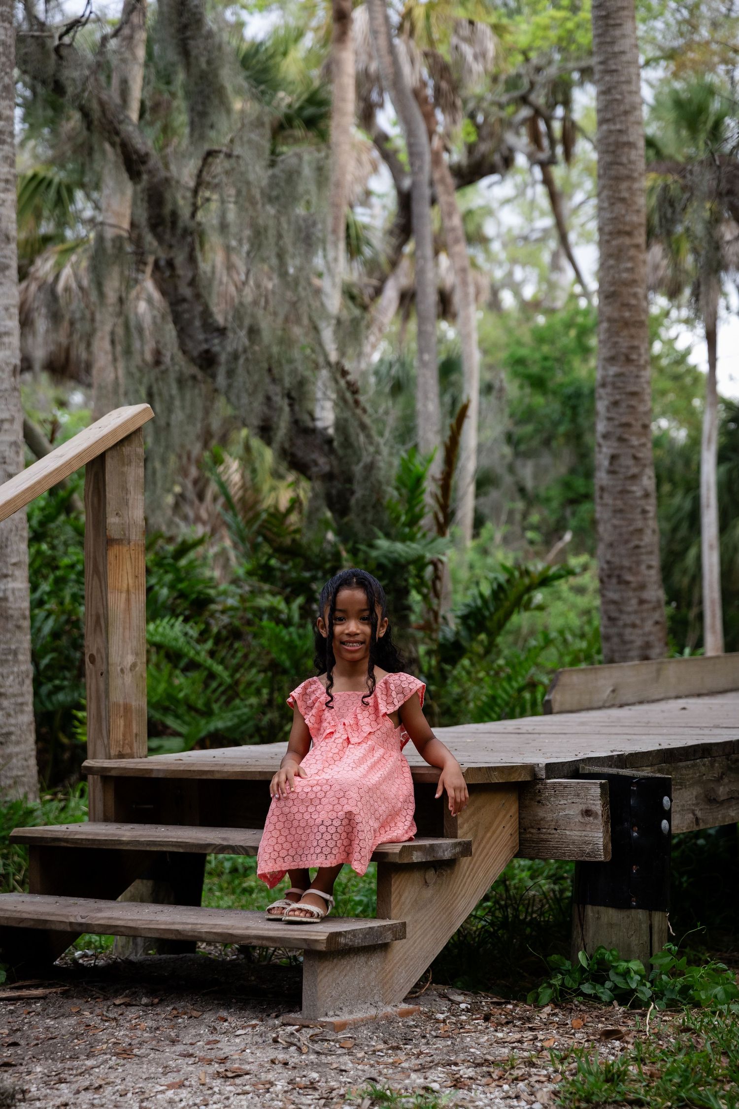 Young girl in pink polka dot dress sitting on wooden steps surrounded by lush green trees and palm trees.