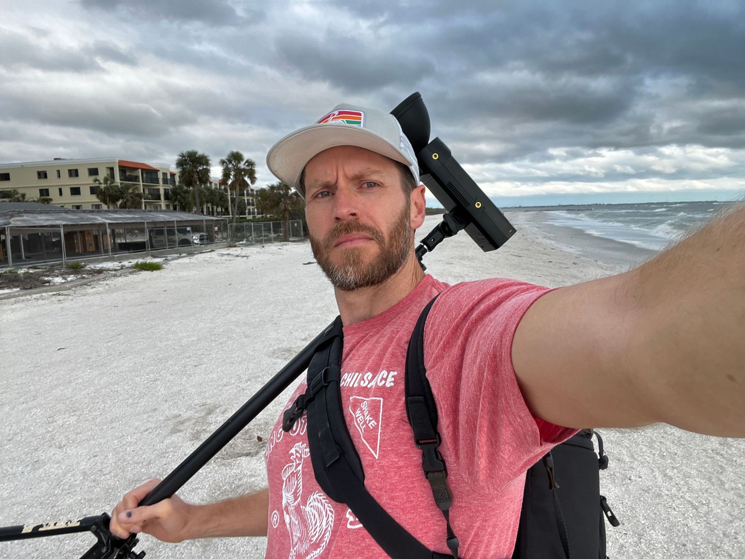 Man taking a selfie on a white sand beach with a camera on a monopod, wearing a pink shirt and cap on a cloudy day.