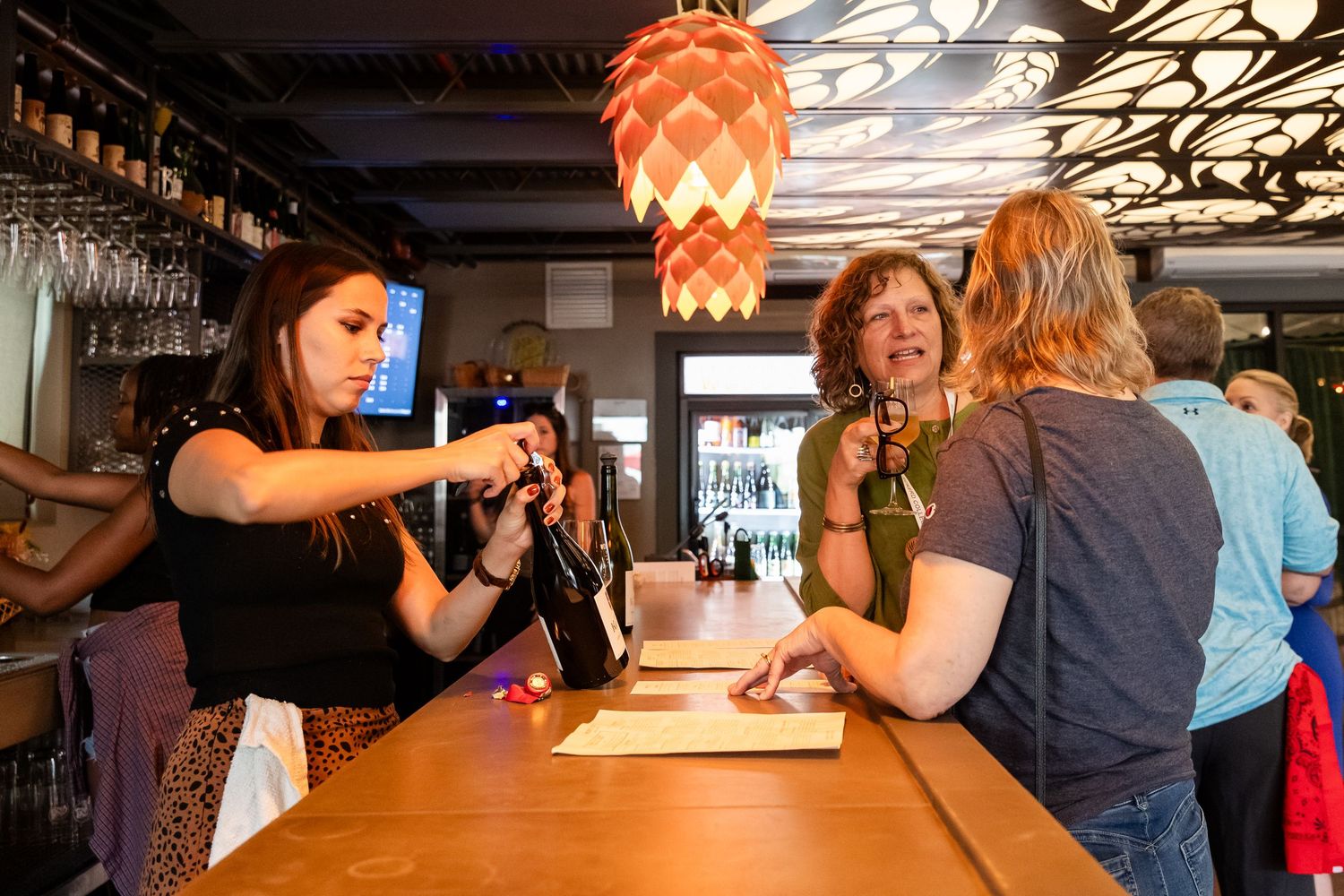 Bartender pouring wine for customers at a modern restaurant bar with hanging pendant lights.