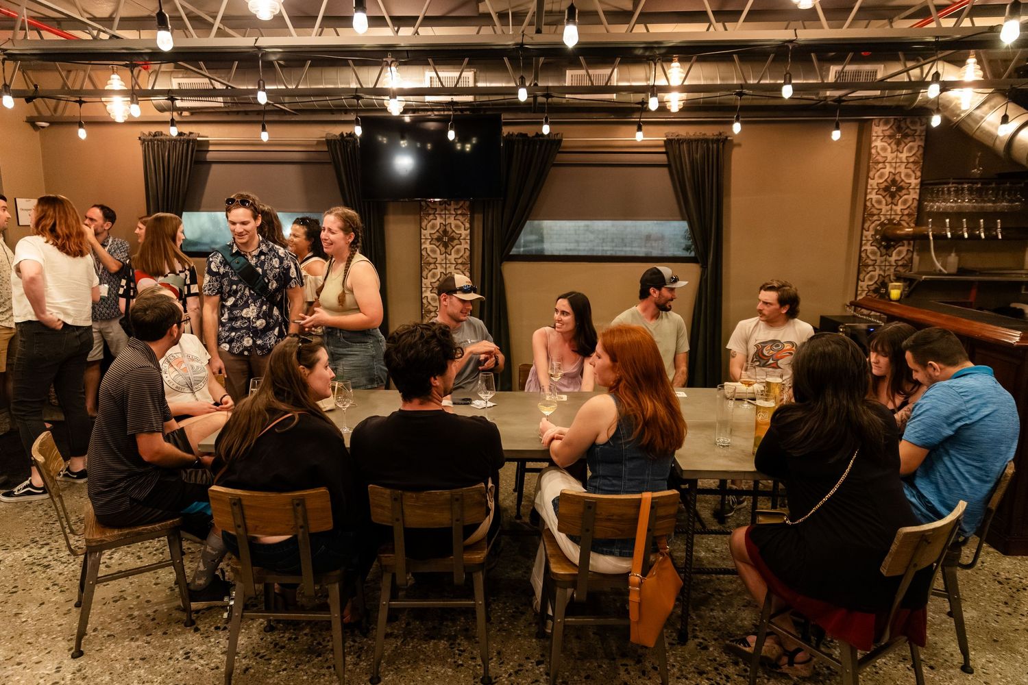 Group of young adults gathered around a large wooden table in a dimly lit brewery taproom during a social event.
