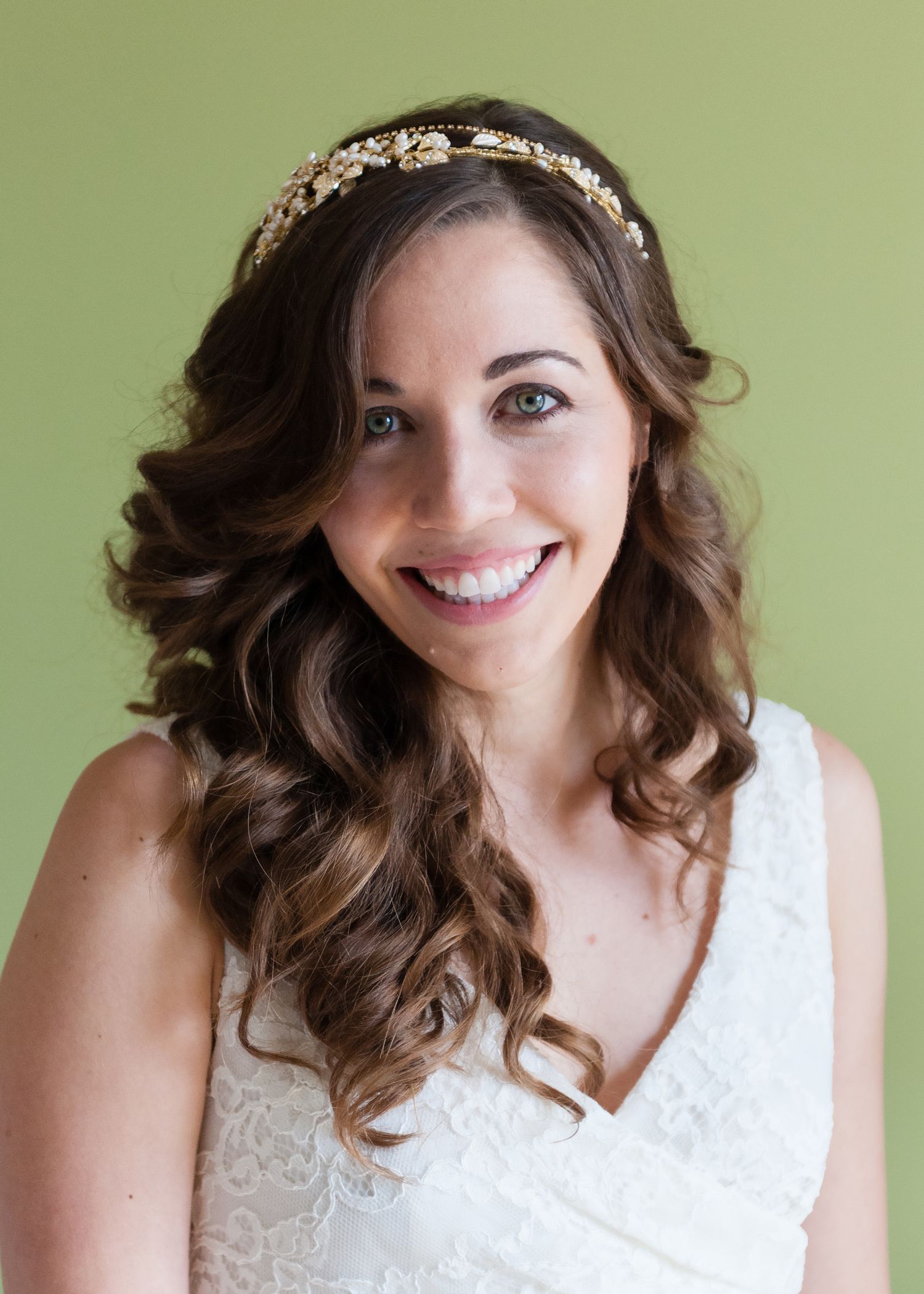 Smiling bride wearing a gold floral headband and white lace dress with curled brown hair.