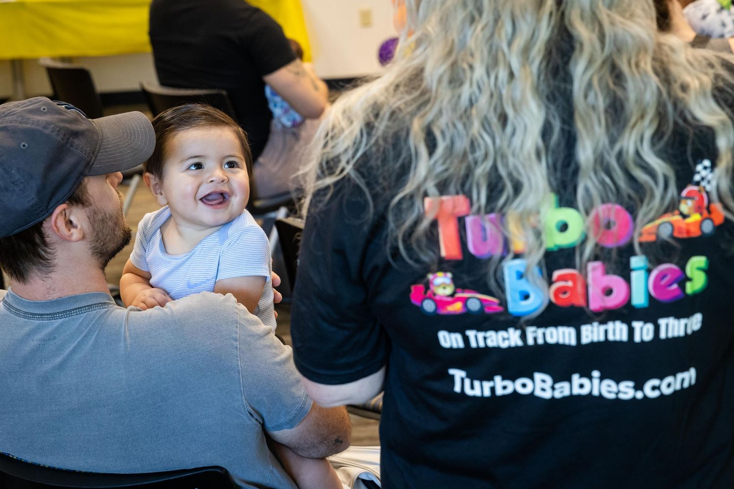 A smiling baby is held by an adult near a Turbo Babies staff member wearing a branded black shirt at a community event.