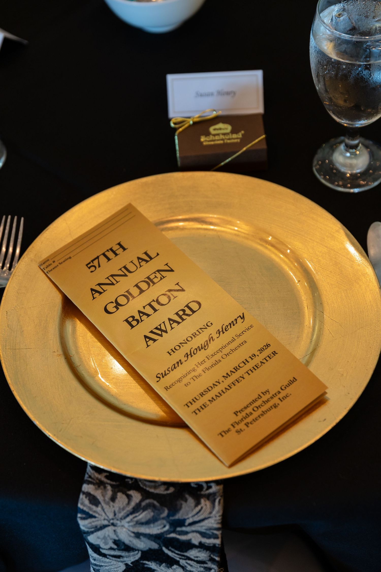 Gold charger plate with 57th Annual Golden Baton Award program at an elegant black-themed banquet table setting.