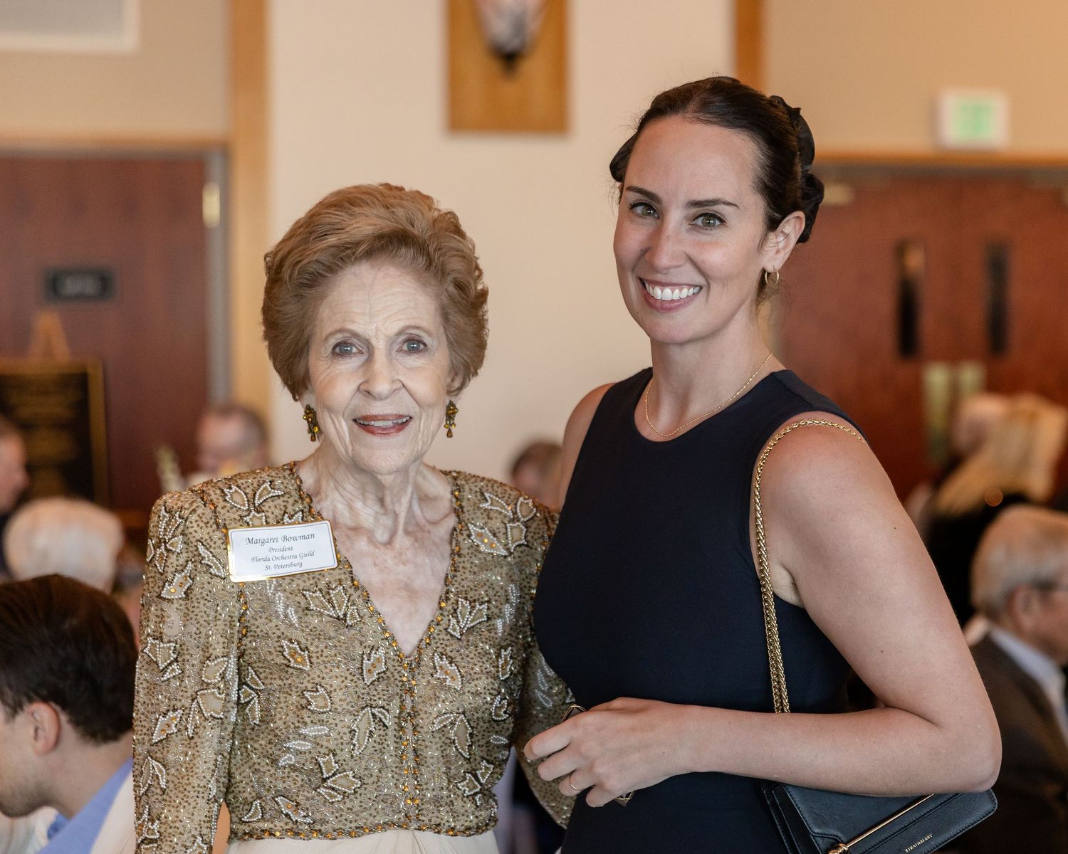 Two women smiling together at formal indoor event, one elderly in gold lace dress.