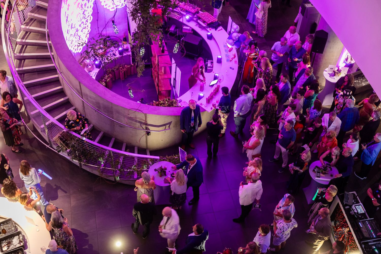 Aerial view of an elegant event venue with purple lighting, spiral staircase, and guests mingling at a upscale party.