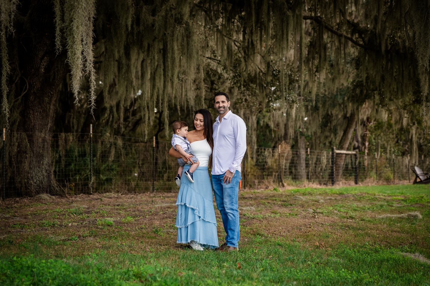 Family of three poses under Spanish moss-draped oak trees on green grass, mother in blue dress holding baby.