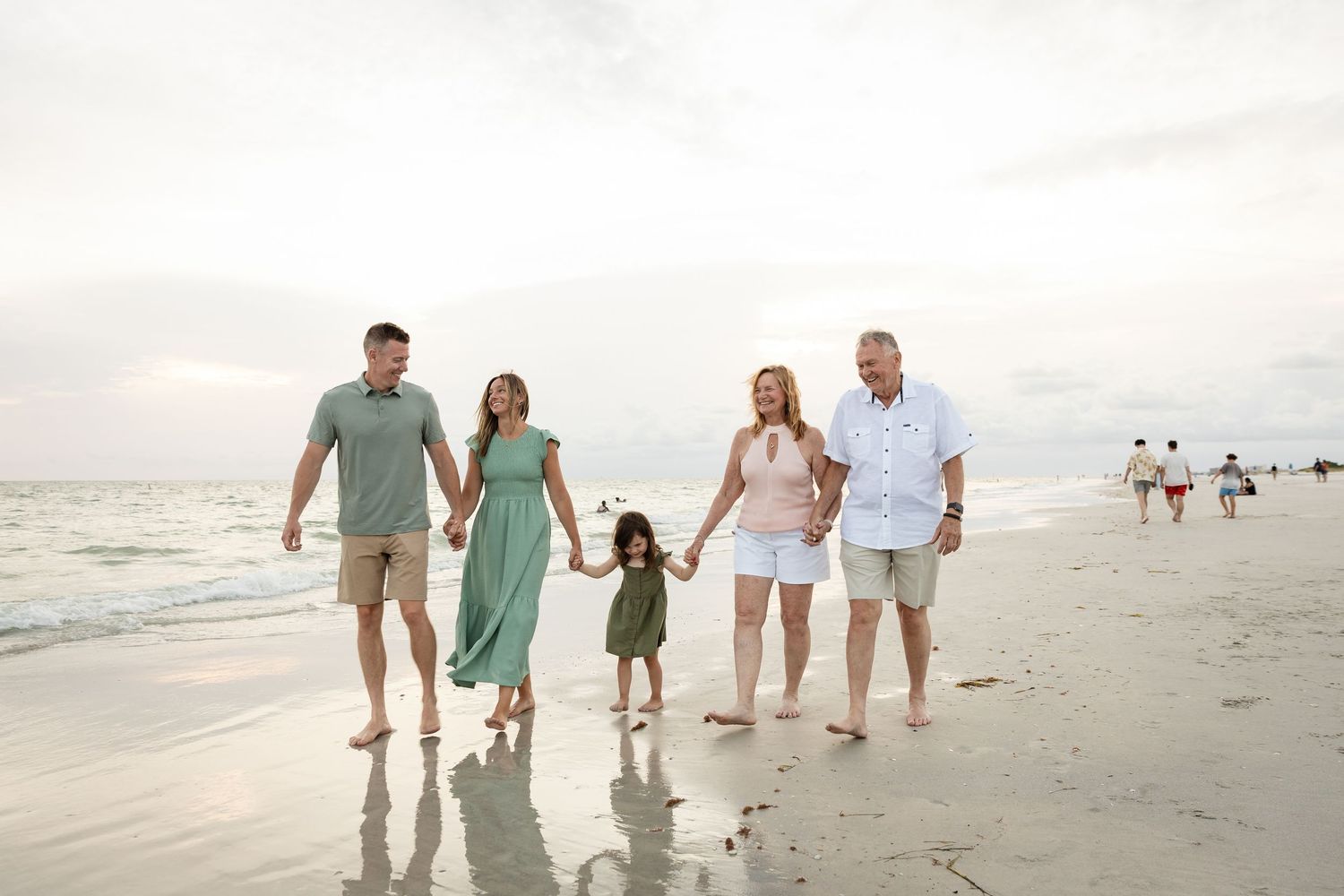 Family of five walking hand-in-hand on a sunny beach, dressed in coordinating green and white outfits.