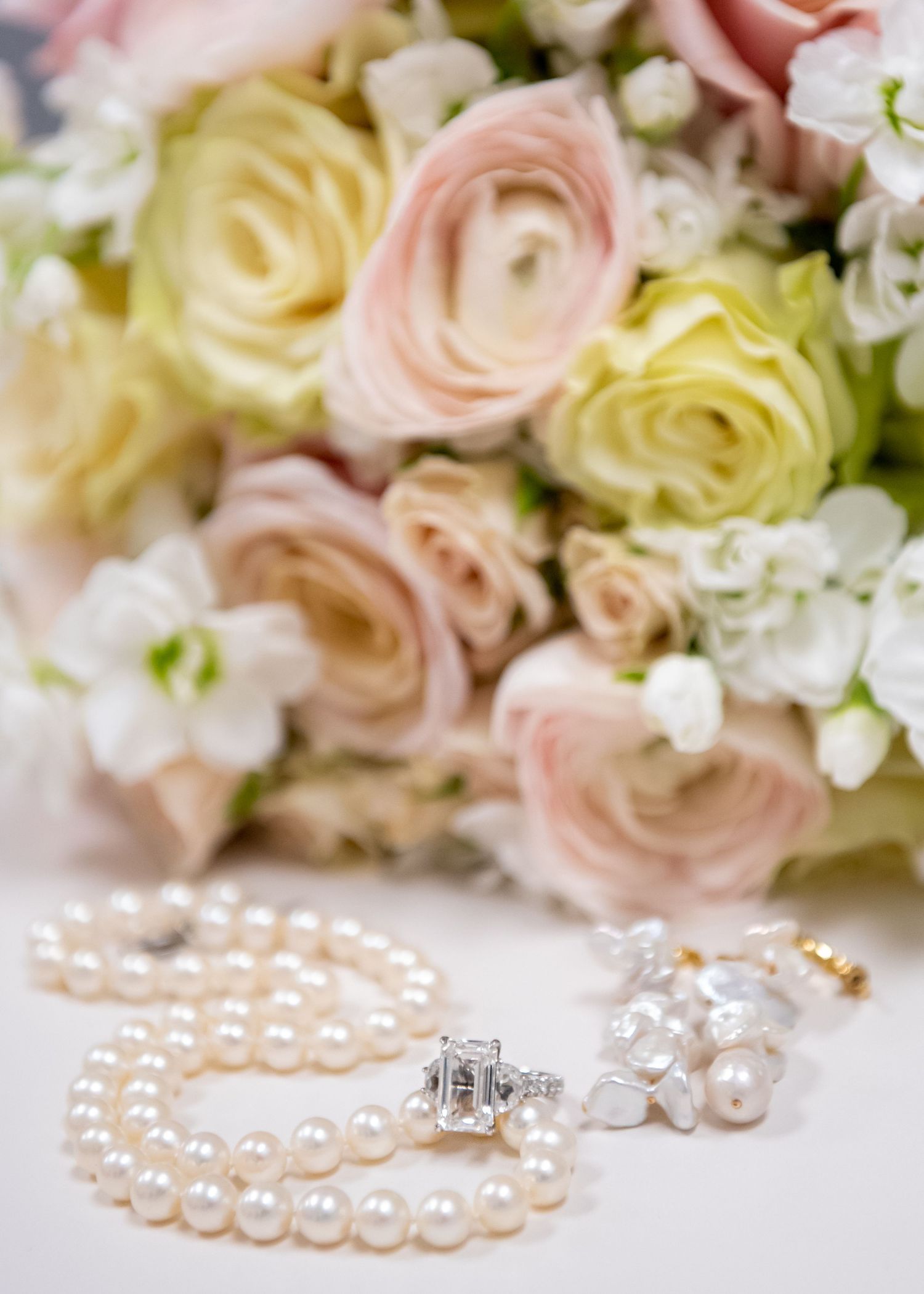 Pearl necklace and diamond ring beside elegant pink and white rose bridal bouquet on white surface.