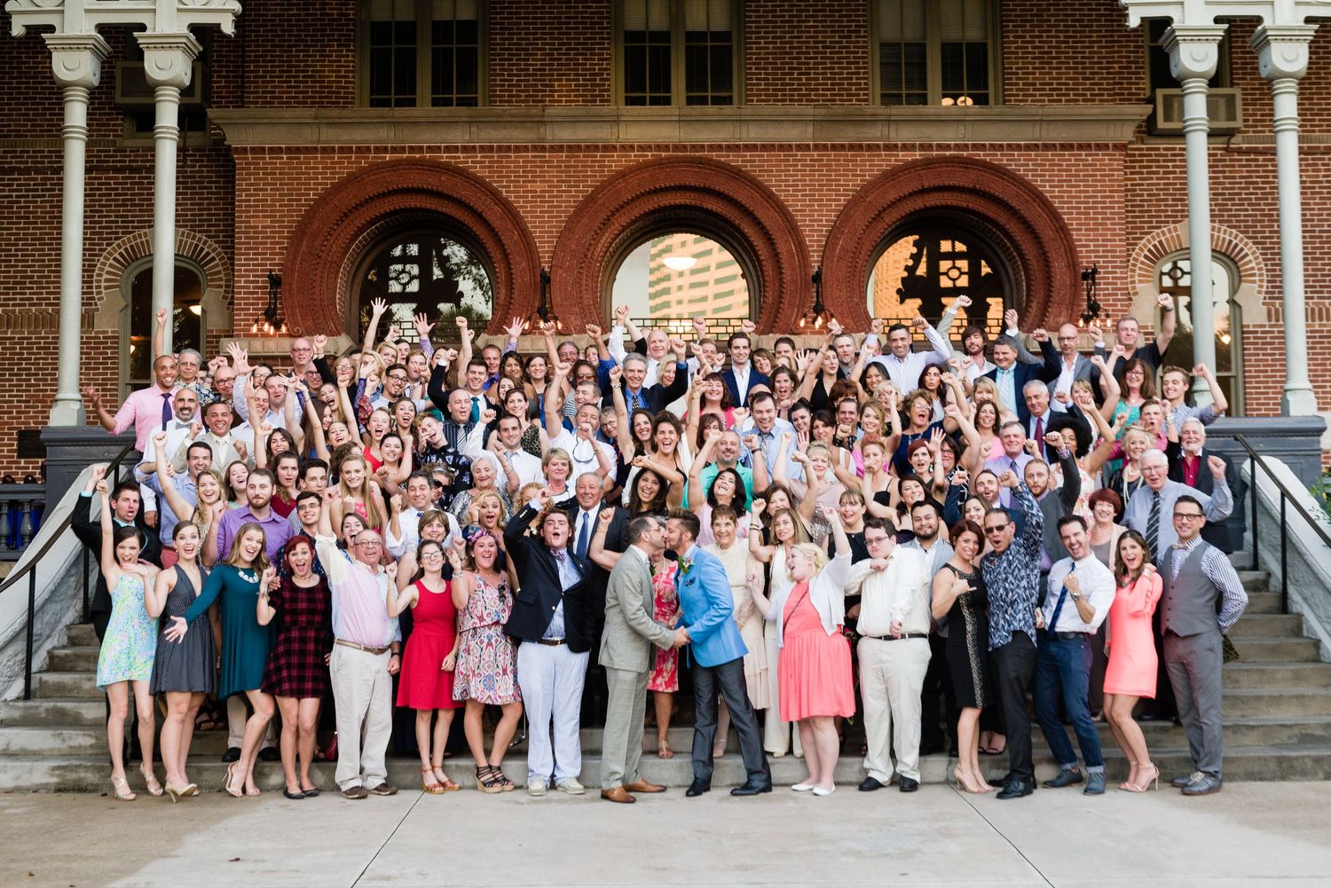 Large group of elegantly dressed people posing on steps of a brick building with arched windows.