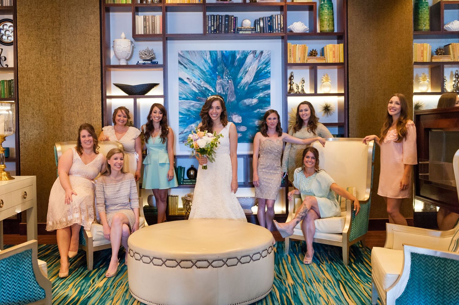 Bride poses with bridesmaids in mismatched pastel dresses inside elegant hotel lobby with teal carpet.