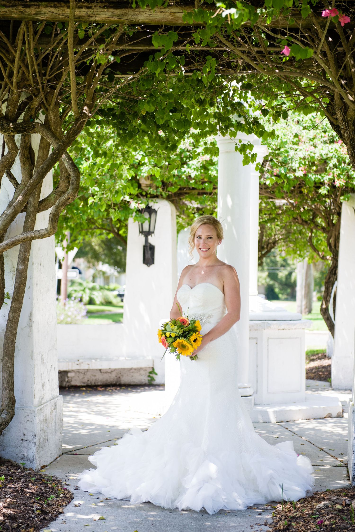 Elegant bride in a strapless mermaid gown holds a sunflower bouquet under a lush green vine-covered pergola outdoors.