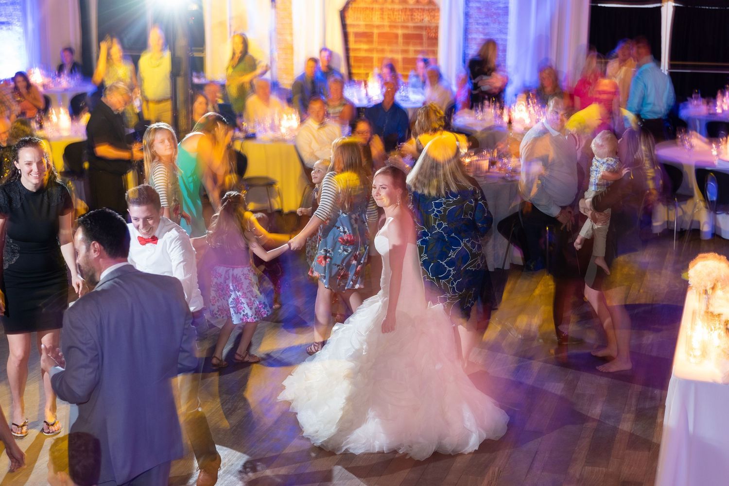 Bride in white gown dancing with guests at elegant wedding reception with candlelit tables.