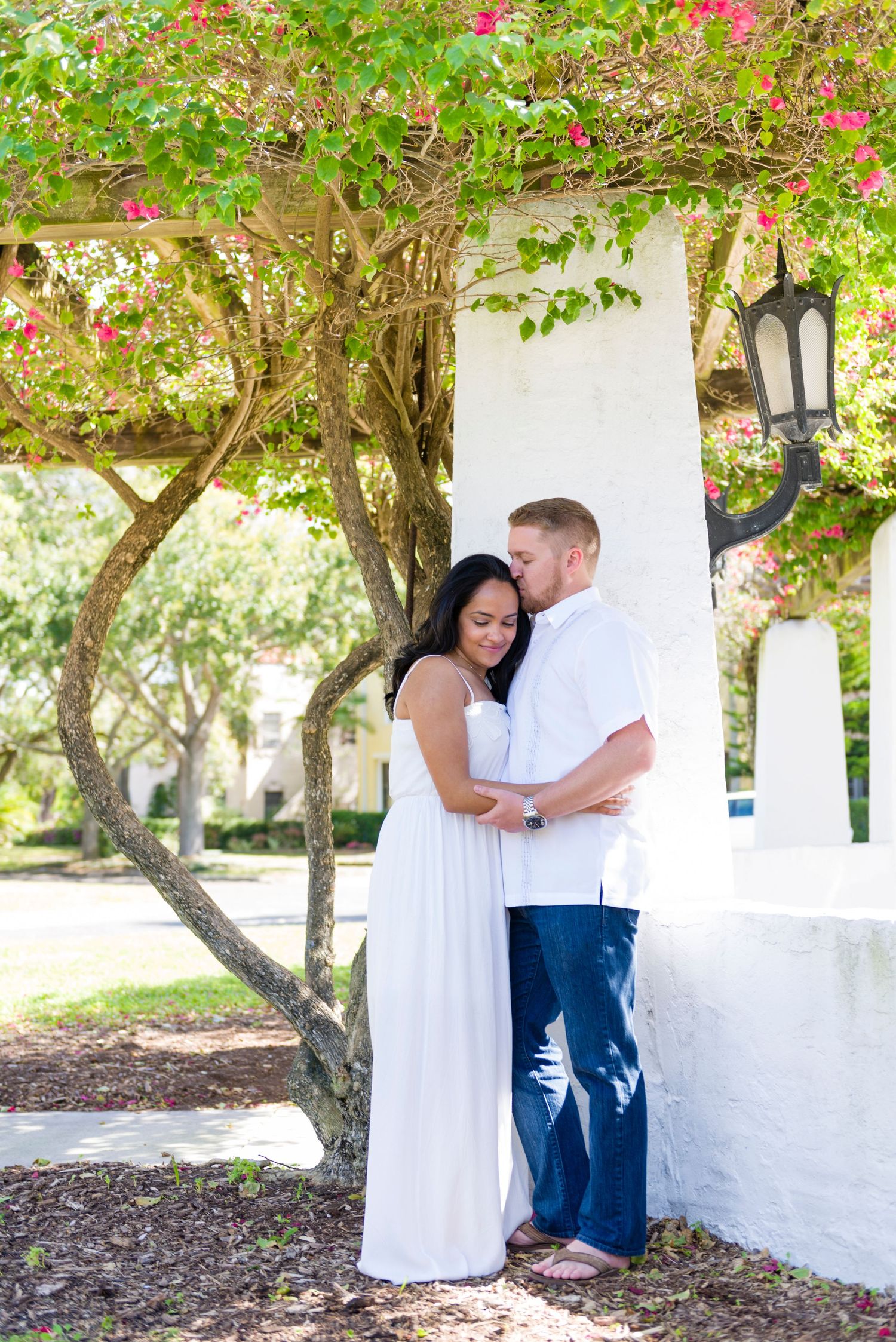 Couple in white outfits embracing under flowering bougainvillea tree by white column in sunny outdoor setting.