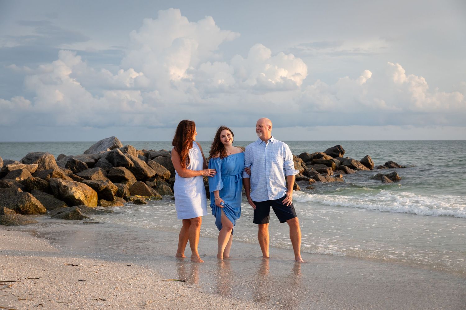 Family of three smiling and posing on a sandy beach with rocky jetty and cloudy sky backdrop.