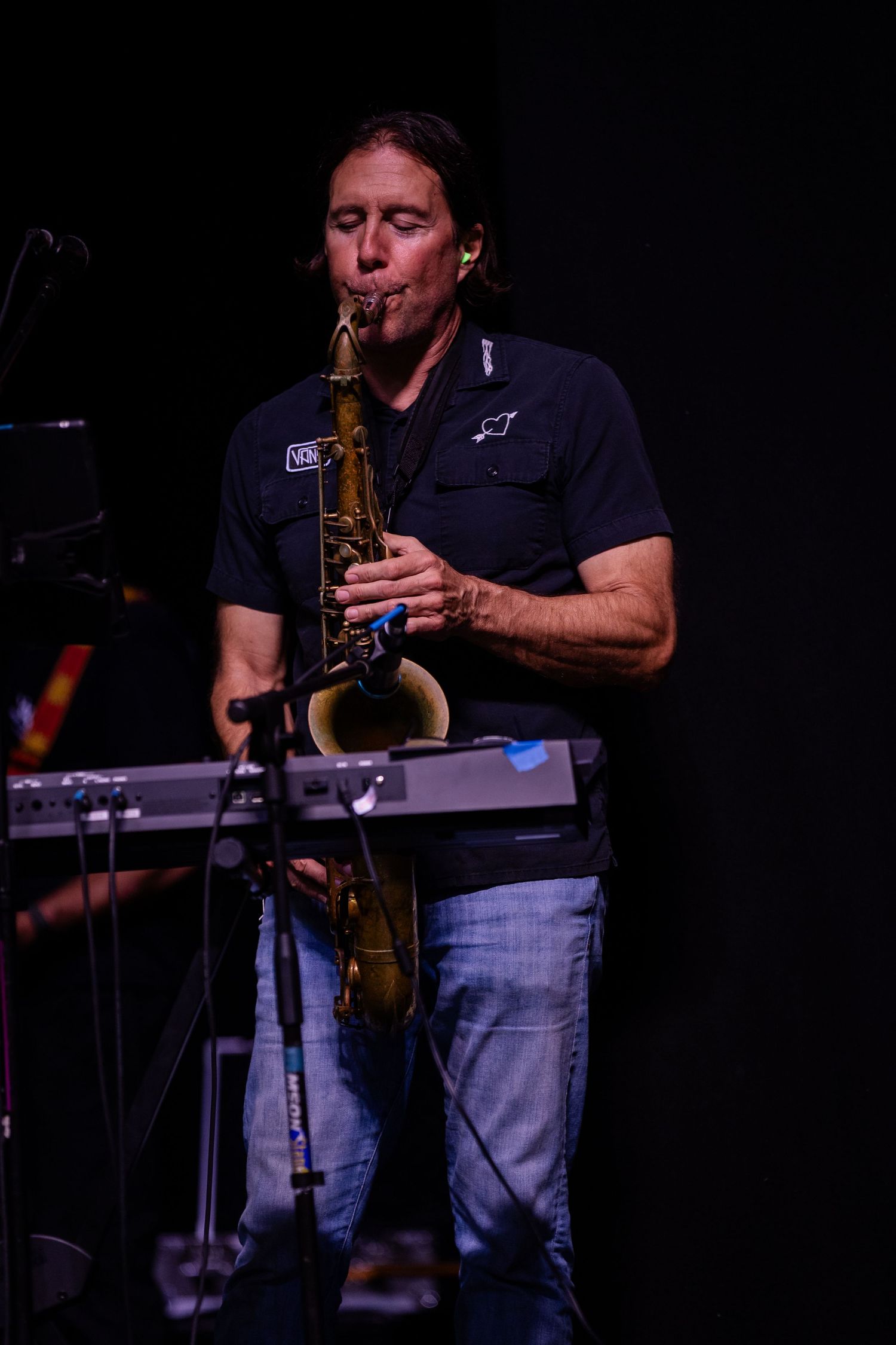 A male musician performs on saxophone on a dark stage wearing a black shirt and jeans.