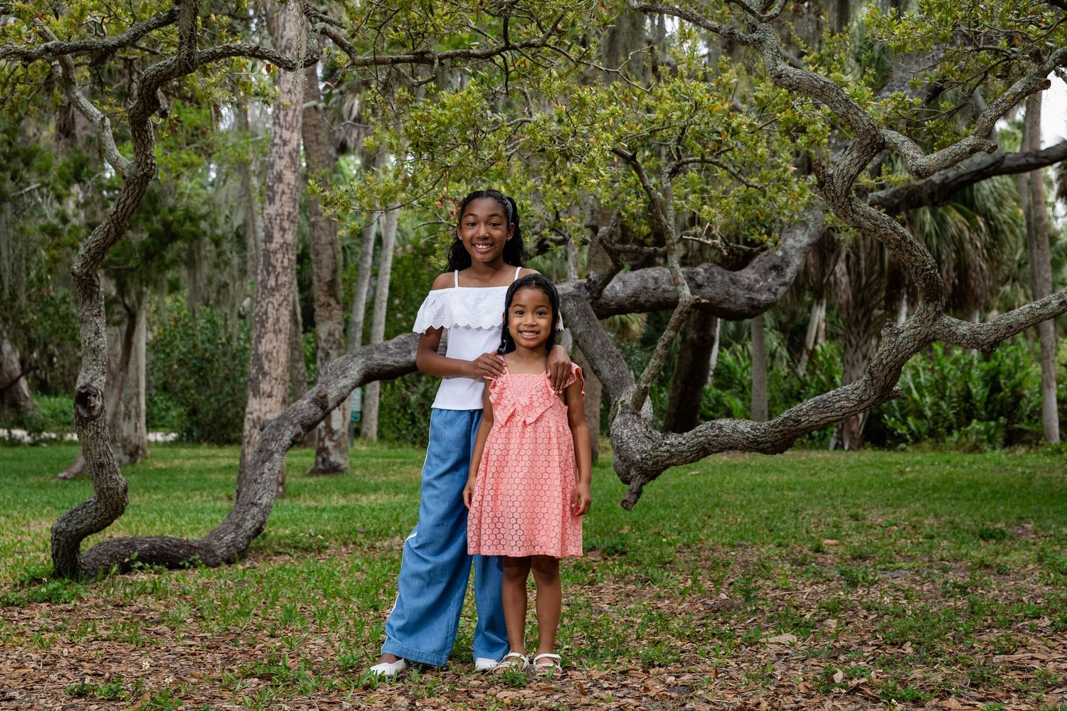 Two young girls pose together in a lush green park surrounded by large trees.
