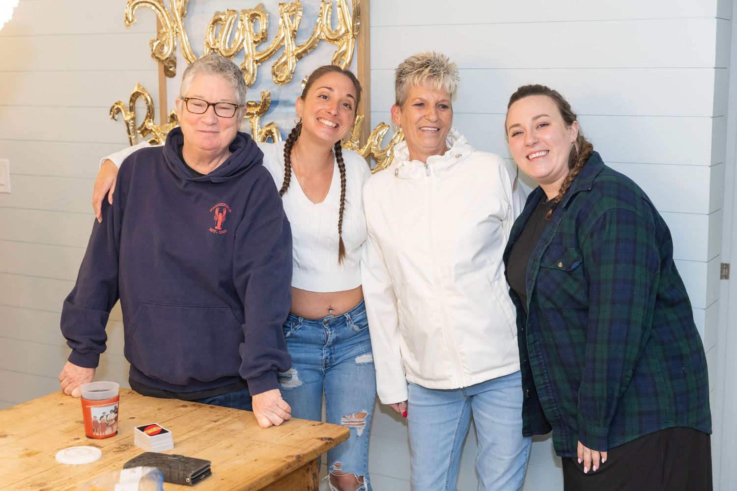 Four people smiling together at a Happy Birthday celebration near a wooden table with gold balloon decorations.