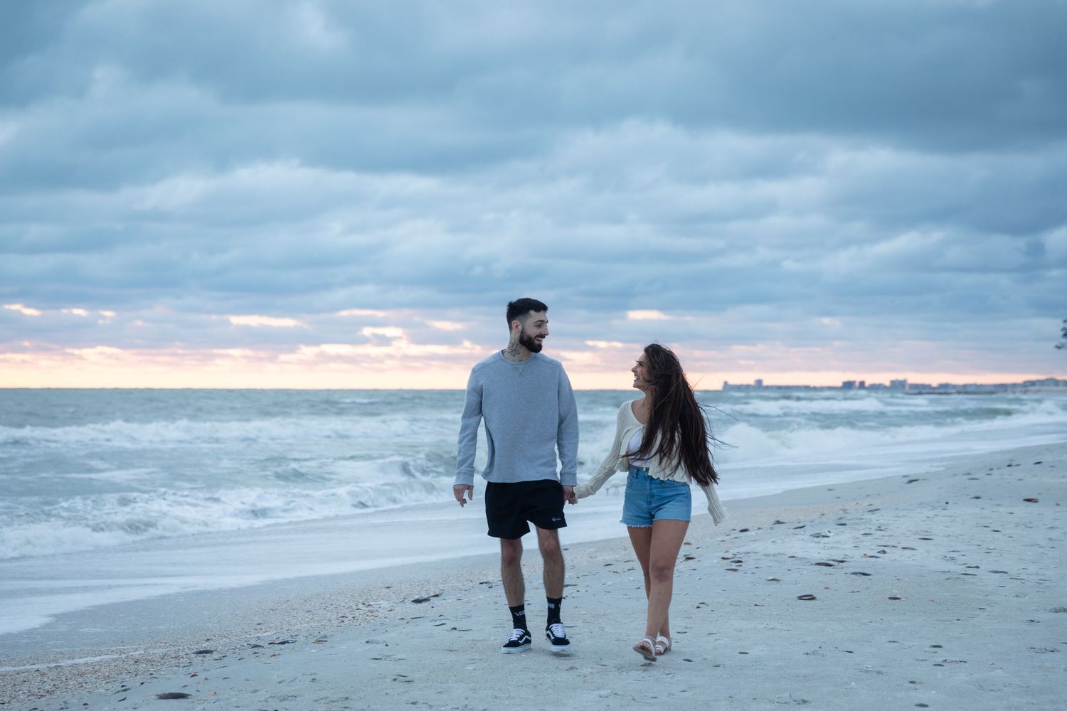 Couple holding hands walking on a sandy beach at sunset with cloudy skies and ocean waves.