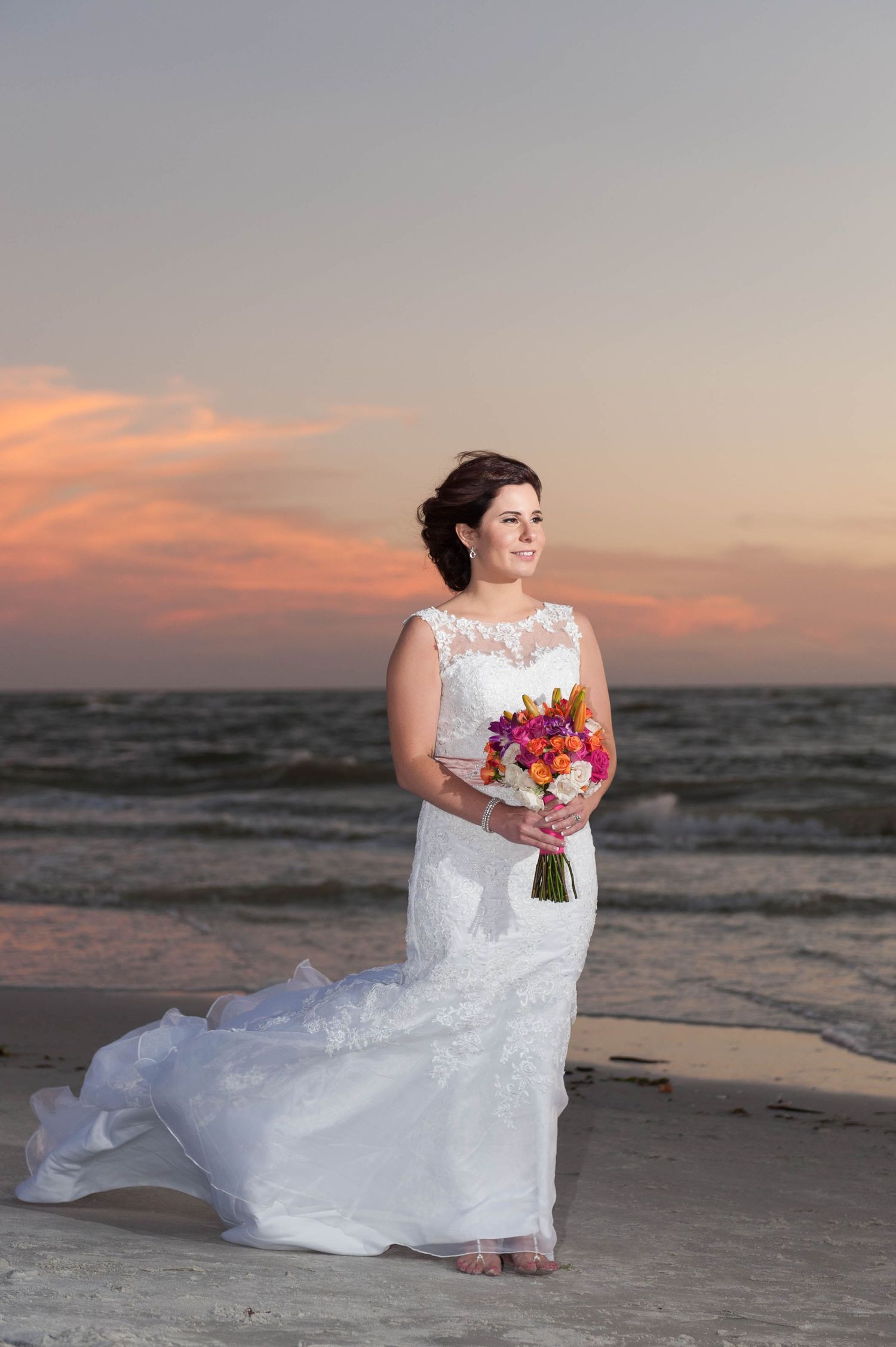 Bride in lace wedding gown holds colorful bouquet on beach at sunset with ocean waves behind her.