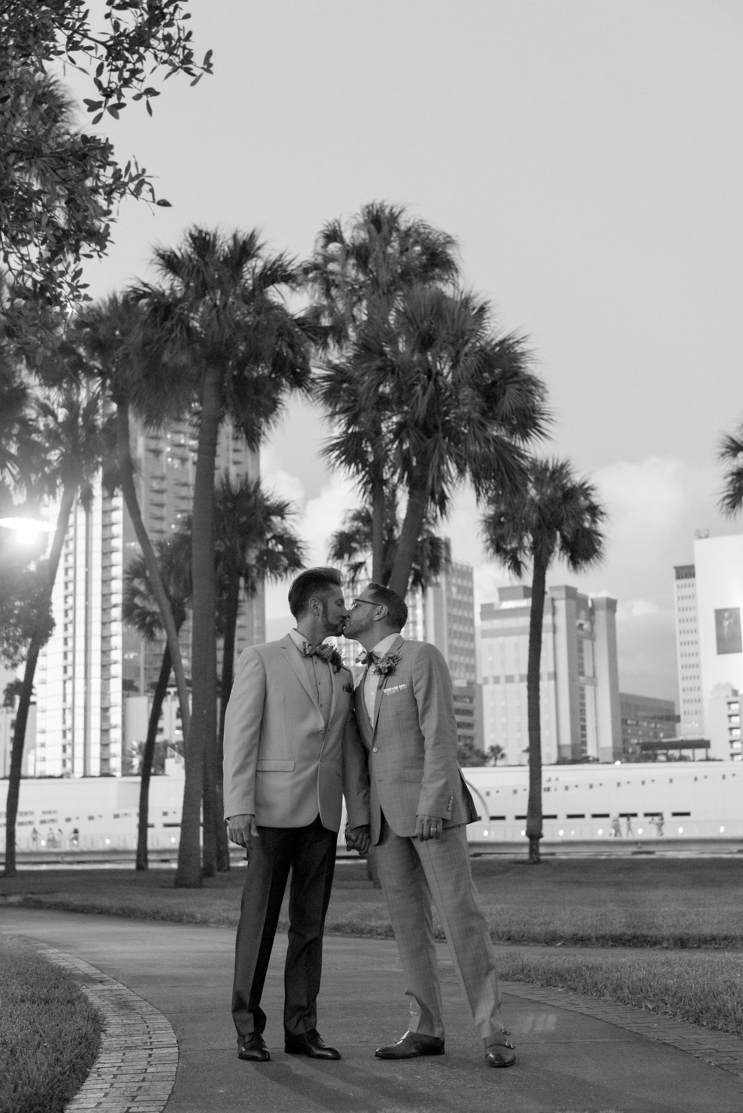 Two grooms share a kiss in black and white wedding photo with palm trees and city skyline backdrop.