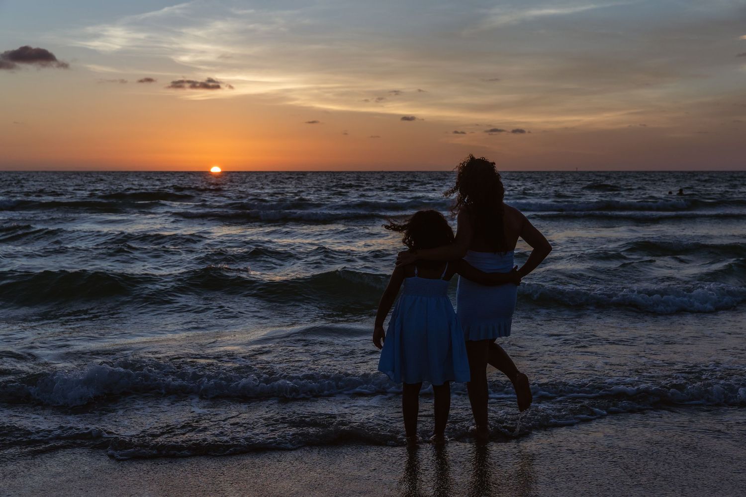 Mother and daughter in blue dresses watch a vibrant sunset over ocean waves on a sandy beach.