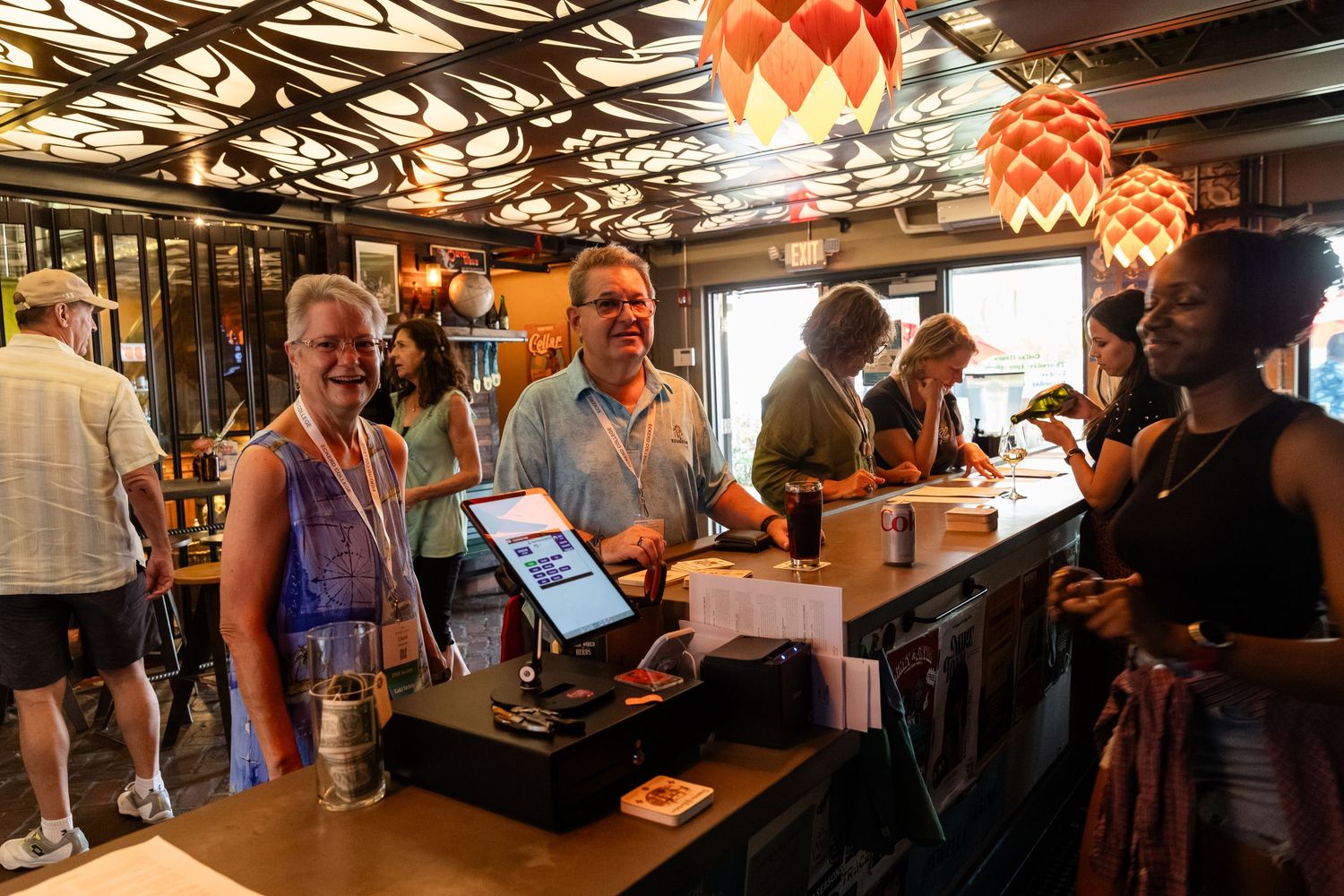 People smiling at a bar counter with colorful hanging lights in a busy restaurant setting.