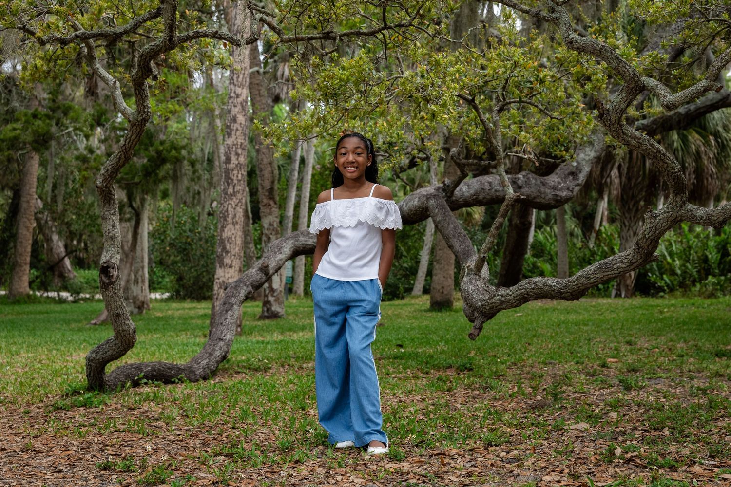 Young girl smiling in white off-shoulder top and blue pants standing among twisted trees in a park.