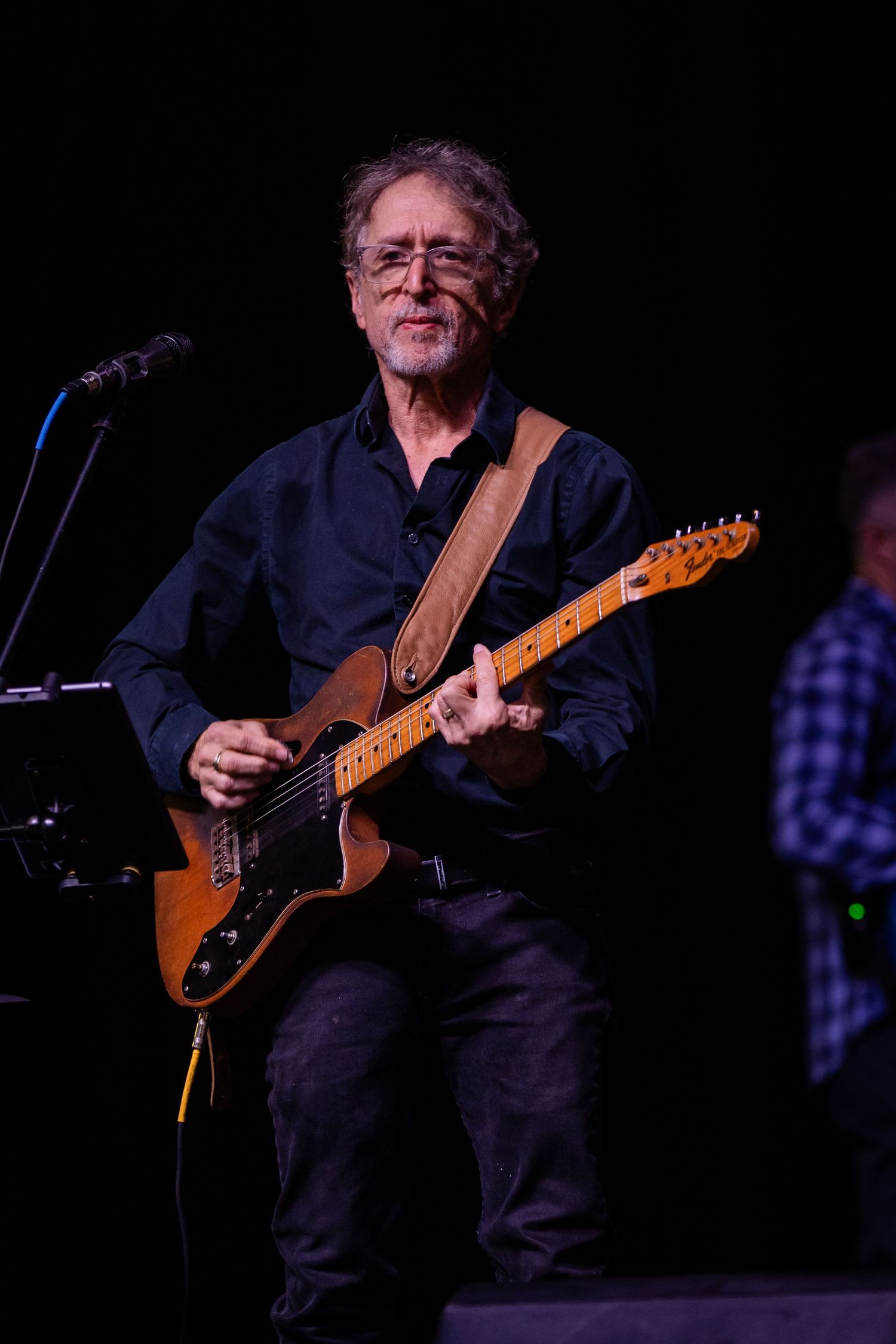 Older man with glasses smiling while playing a brown electric guitar on stage in a dark concert setting.