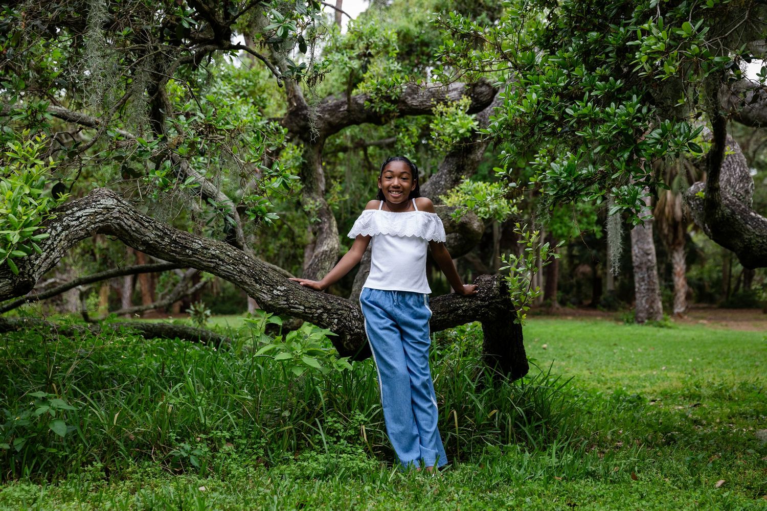 Young woman smiling in lush green park, leaning against a large mossy tree trunk wearing white top and blue pants.