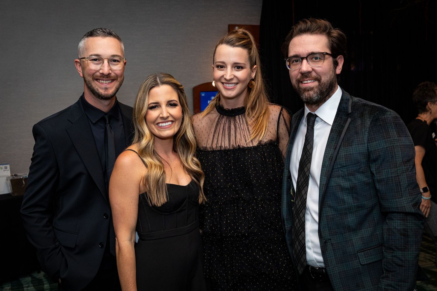 Two couples pose together smiling at a formal evening event, dressed in elegant black and dark attire.