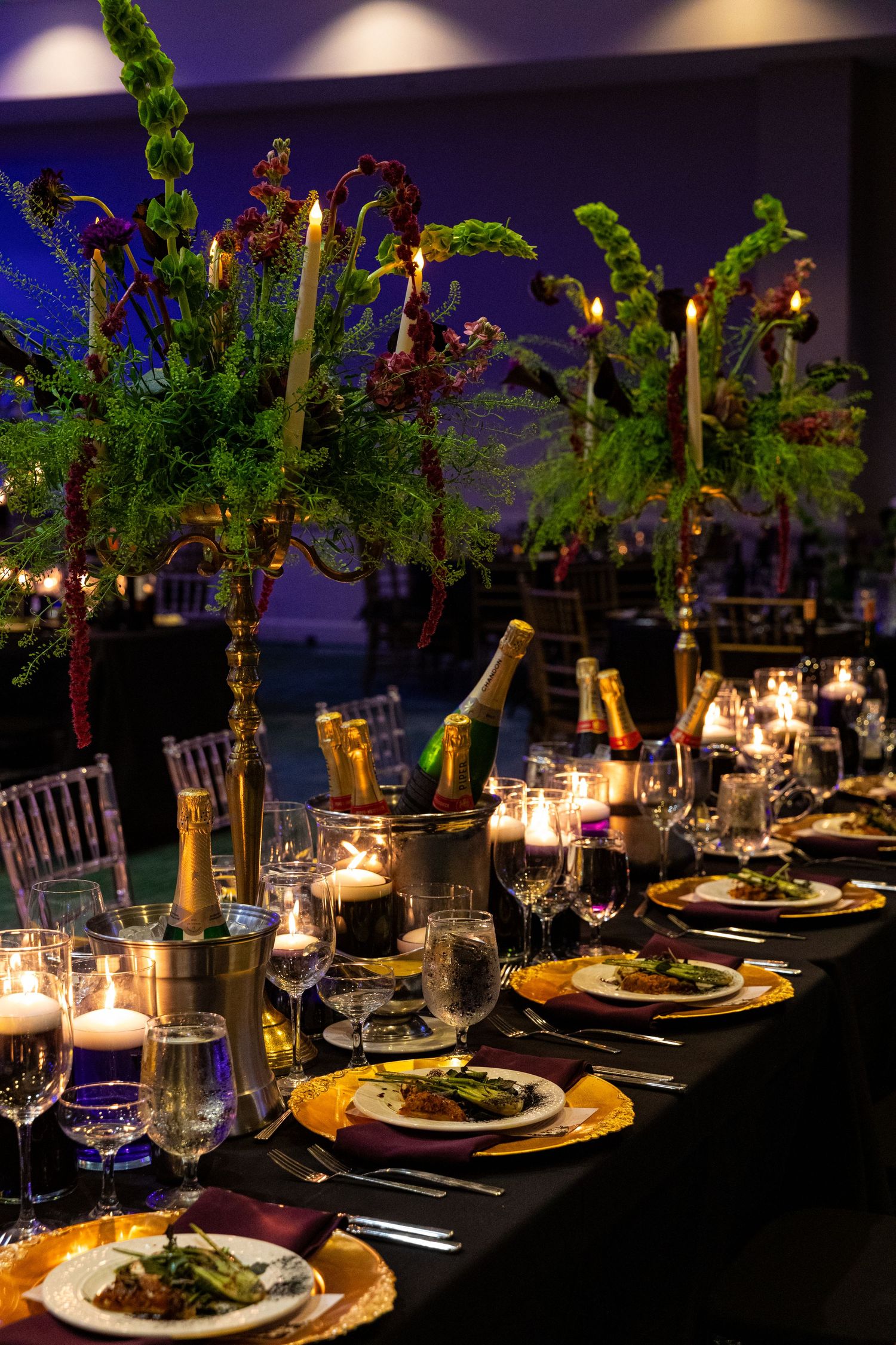 Elegant candlelit banquet table with tall floral centerpieces, champagne, gold chargers, and plated dishes at a formal event.