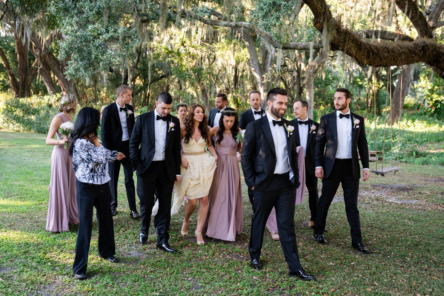 Wedding party in formal attire walks outdoors under moss-draped trees, bridesmaids in pink gowns and groomsmen in black tuxedos.