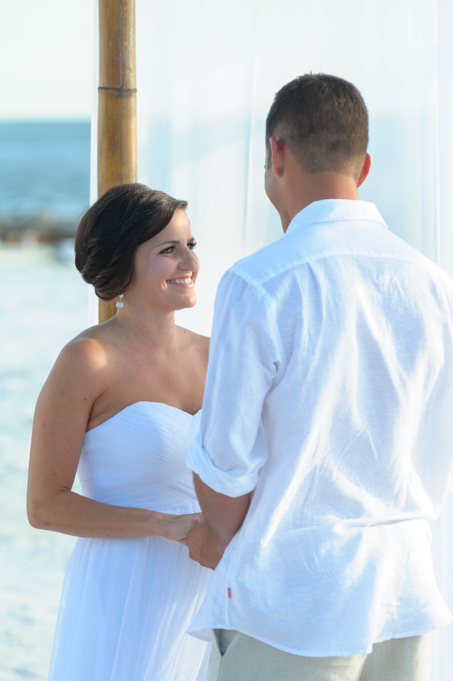 Bride in white strapless gown holds hands with groom during romantic beach wedding ceremony.