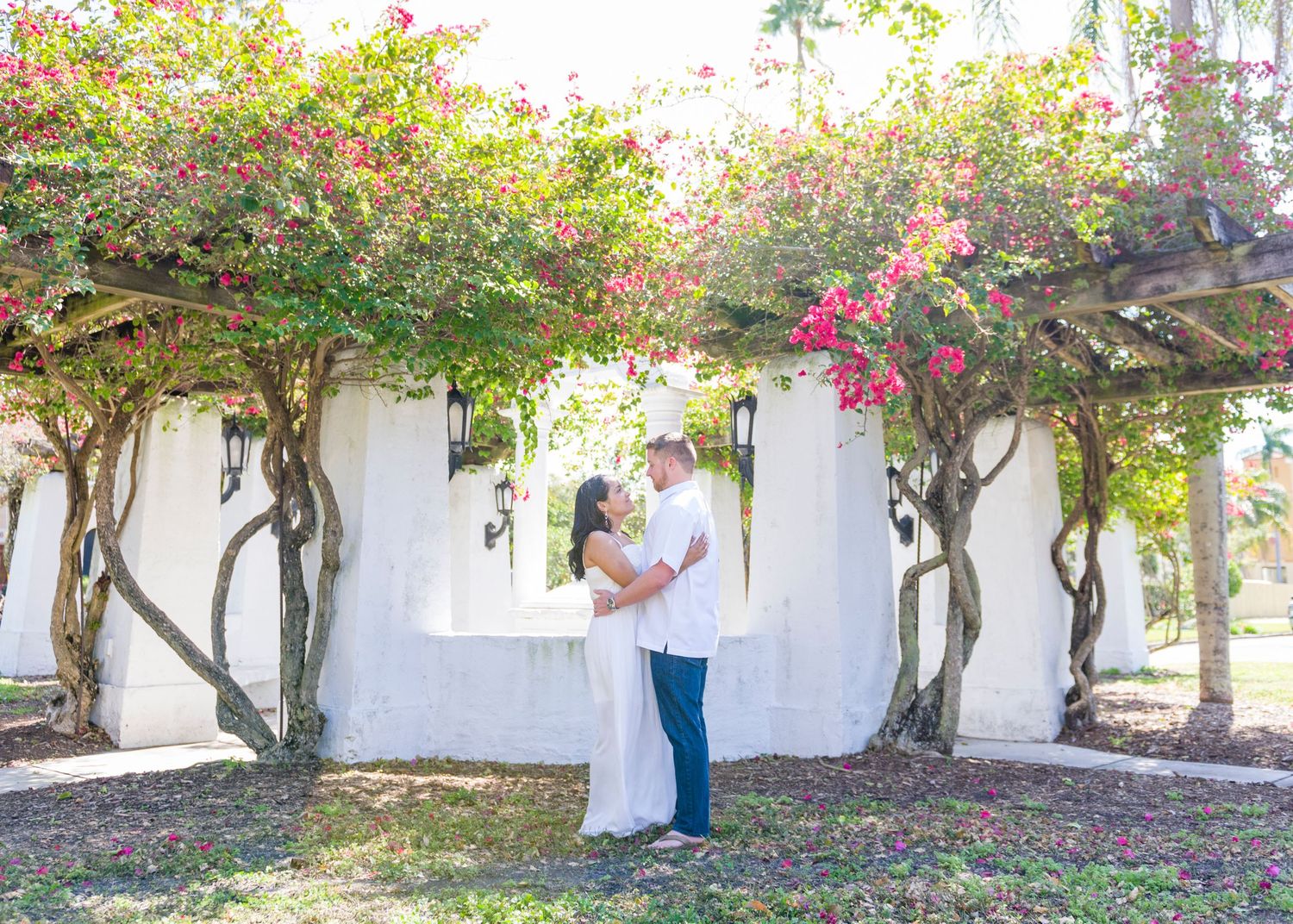 Couple embraces under a flowering bougainvillea pergola with white walls and pink blooms in a romantic outdoor setting.