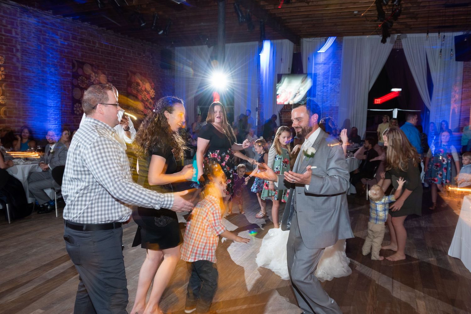 Guests dance and celebrate joyfully at a wedding reception in a warmly lit venue with blue ambient lighting.