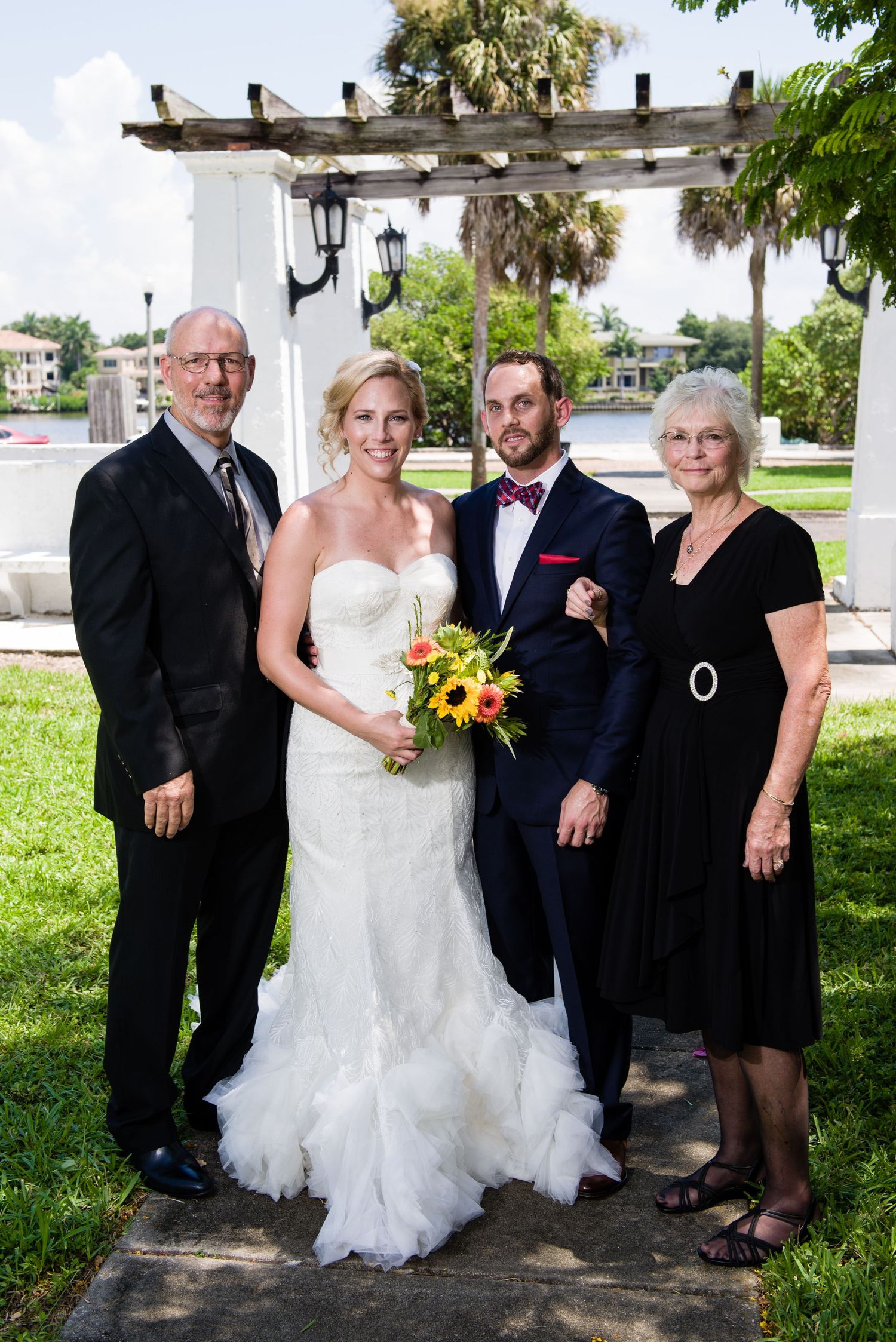 Wedding party of four posing outdoors, bride in white gown with bouquet, groom in navy suit, flanked by family members.
