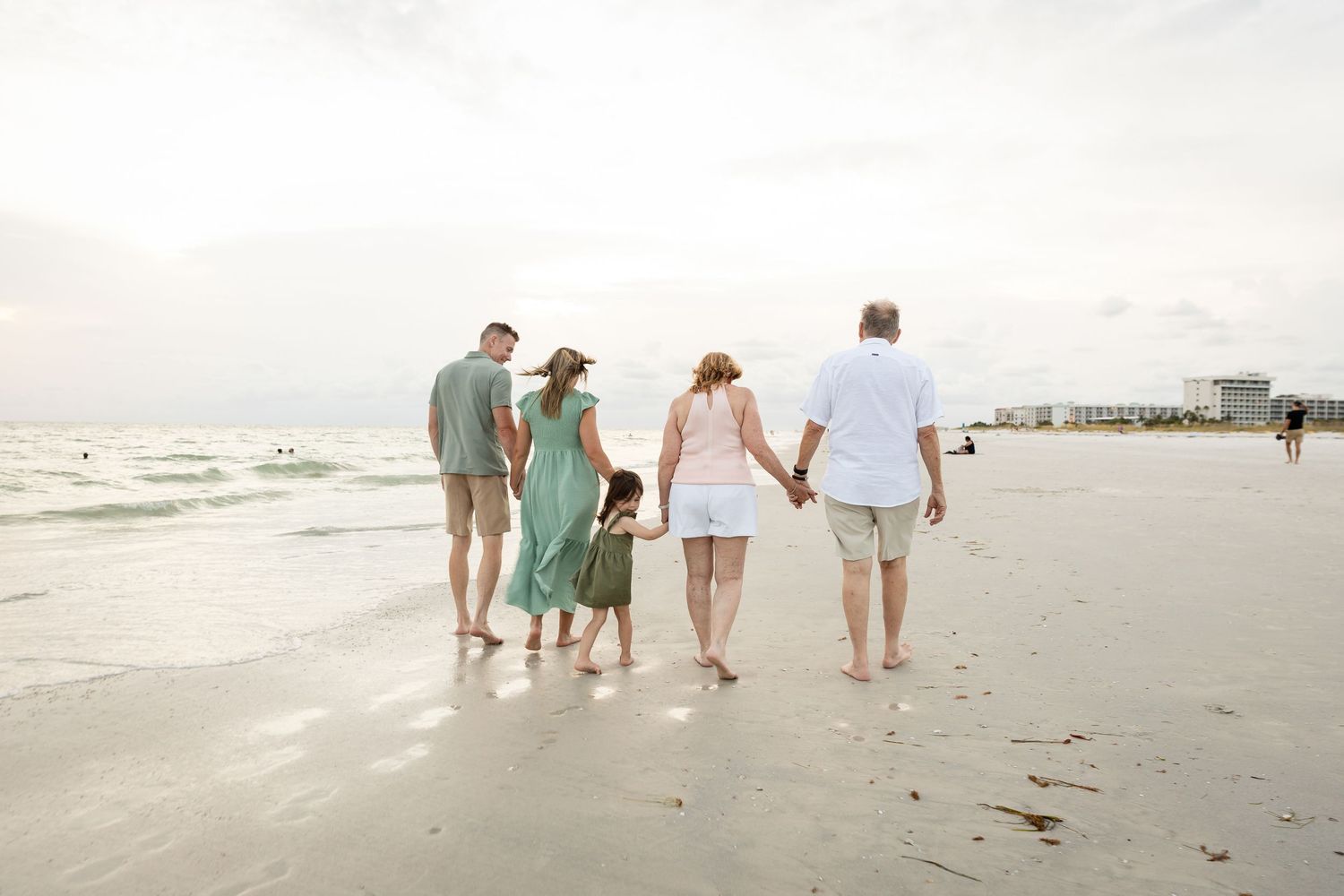 Family of four walking hand-in-hand along a serene sandy beach with calm ocean waves in the background.