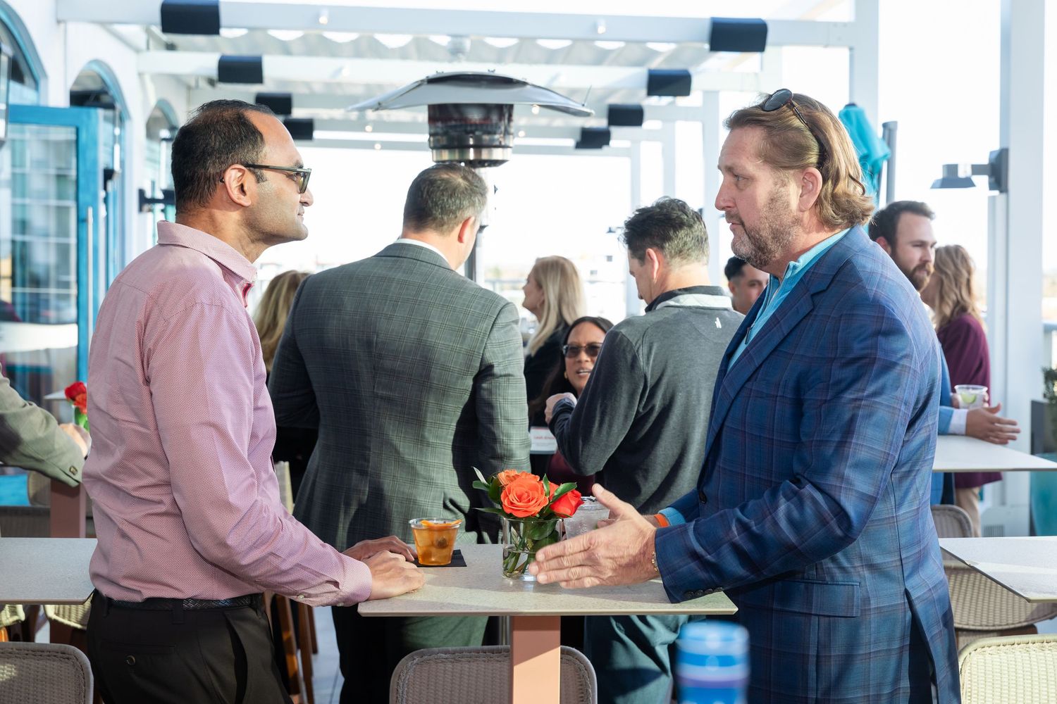 Business professionals networking at an outdoor rooftop event, engaged in conversation around tables with floral arrangements.