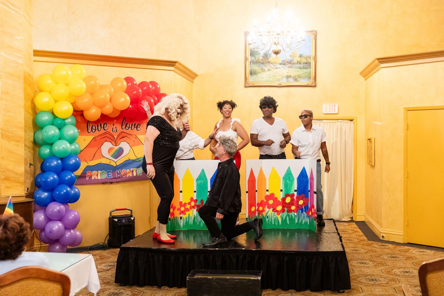 Performers on stage at a colorful Pride event with rainbow balloon arch and Love is Love backdrop decoration.
