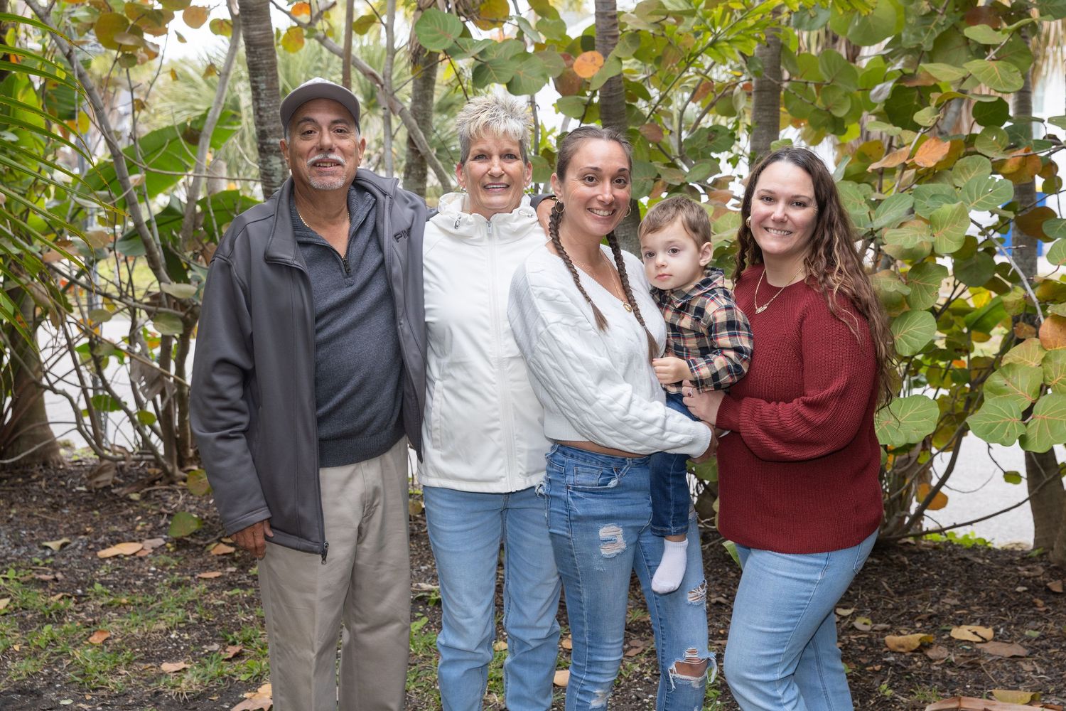 Family of five poses outdoors among tropical foliage, including grandparents, parents, and a toddler.