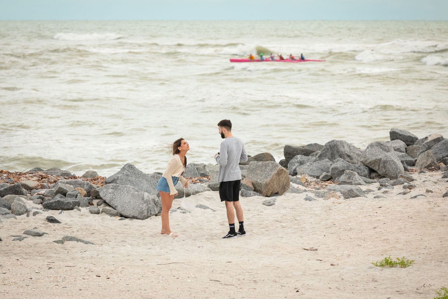 Couple talking on a rocky beach shoreline with a red kayak paddling in the choppy ocean waves behind them.