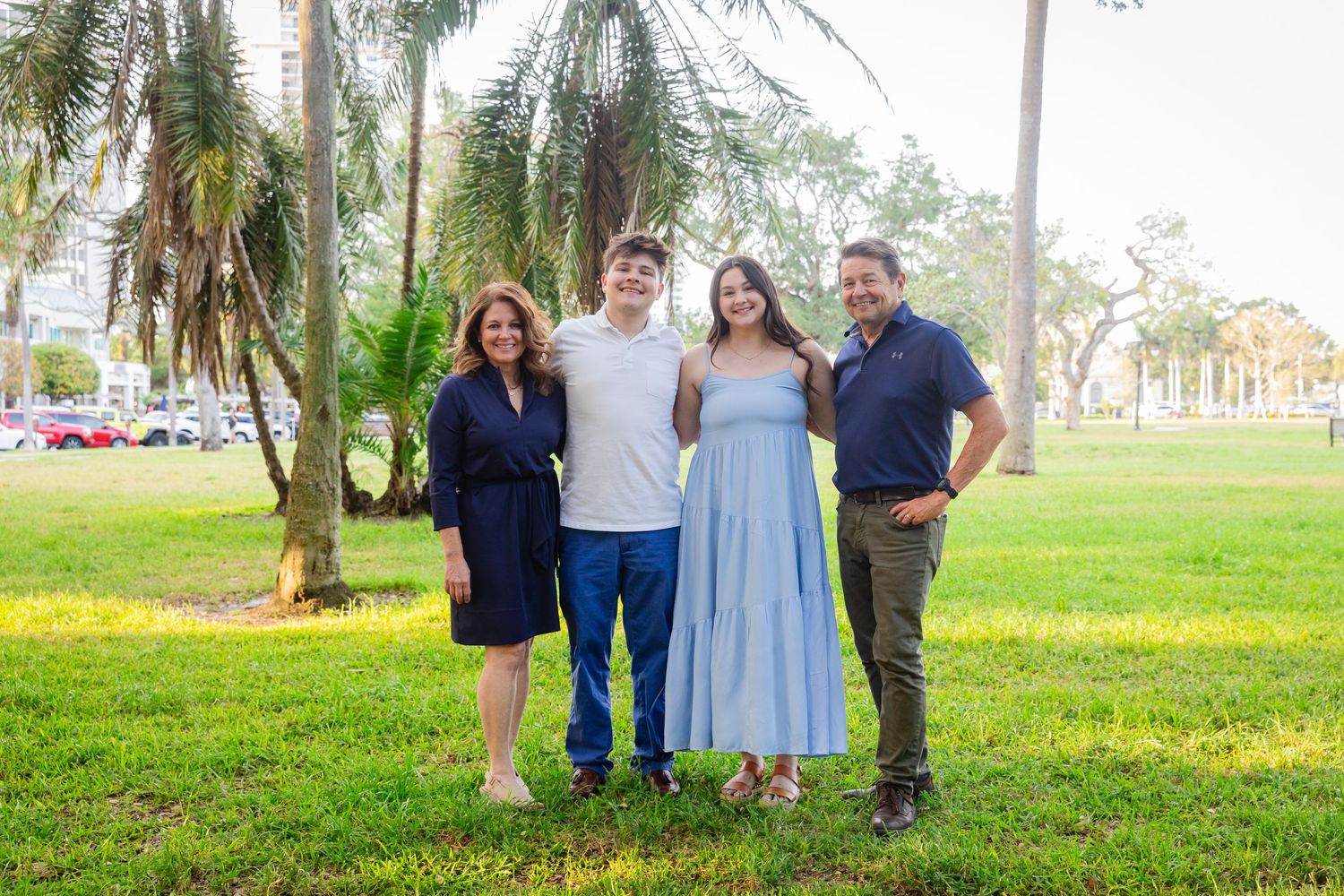 A smiling family of four poses together on a lush green park lawn with tall palm trees in the background.
